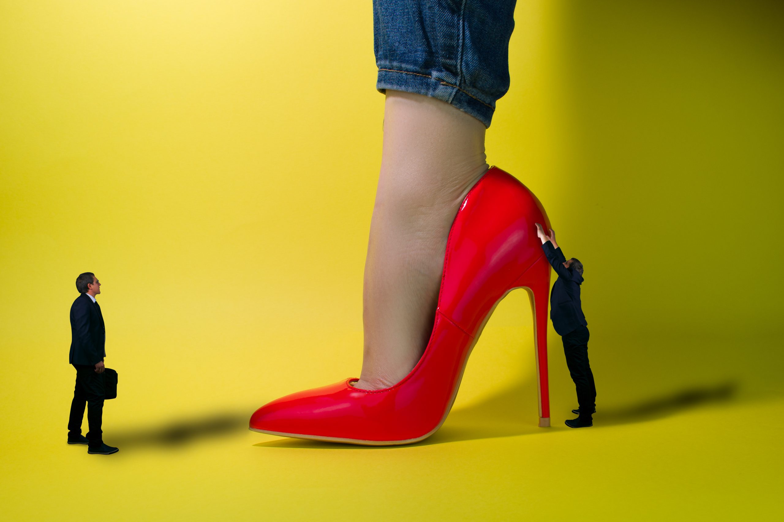 The red high heeled pump of a sexy Asian giantess stands in front of a yellow background while two miniature men in black suits grovel at her feet.