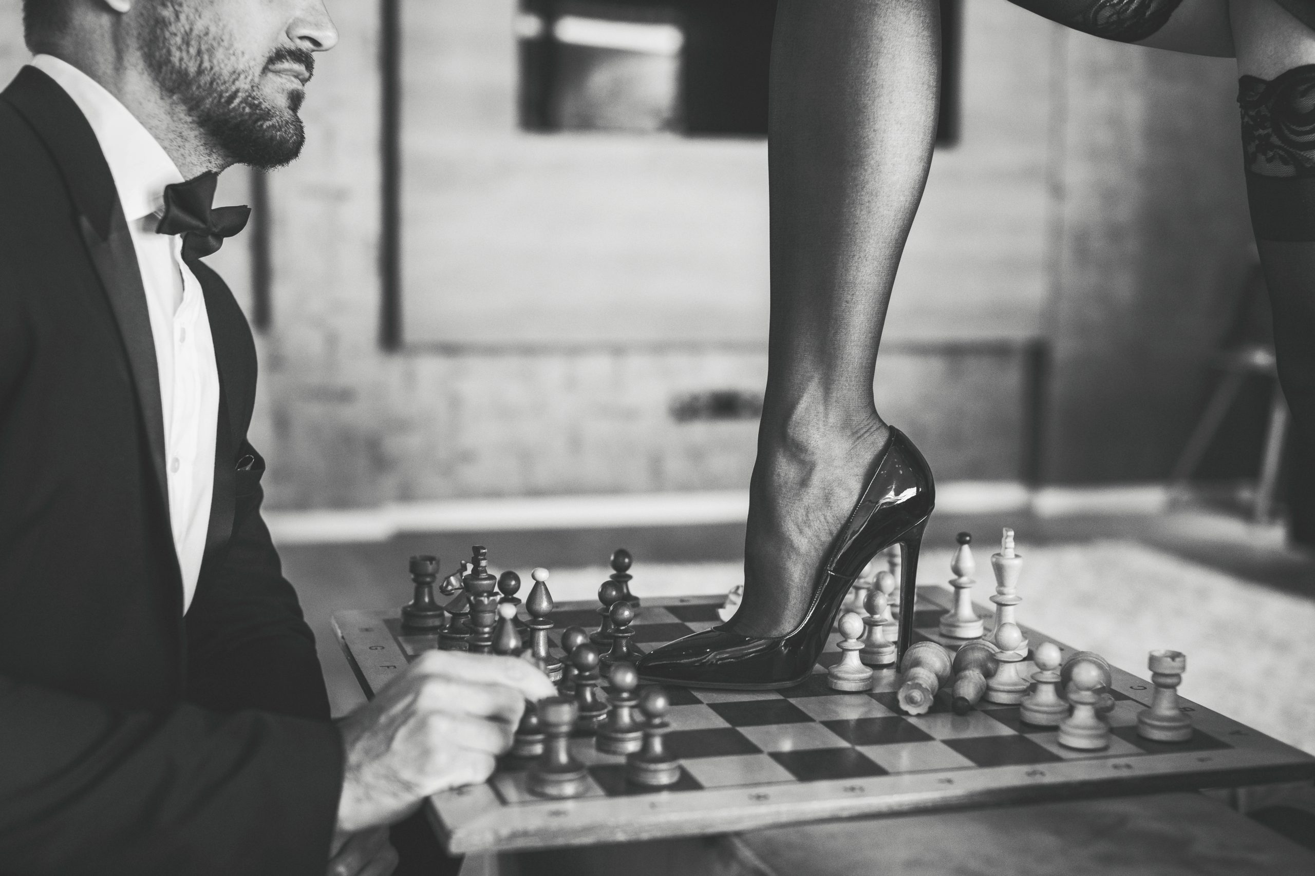 Black and white photo of a financial domination goddess steps her stiletto heel on a game of chess with a paypig dressed in a tuxedo.